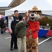 Safety Bear, wearing a red serge standing next to a male Mountie. Safety Bear gives high five to a member of the public.