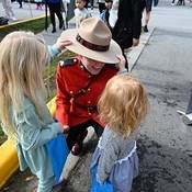 A Mountie in red serge kneeing down while two little girl’s play with the Mountie's Stetson