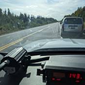A grey minivan is pulled over on the side of a road. A police vehicle’s radar is pictured in the foreground displaying a speed of 143 km/h.