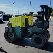 A yellow and green Ammann steamroller, viewed from the side, is stopped next to an orange traffic pylon in a parking lot. No one is on the steamroller and vehicles are visible in the background.