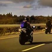 A white motorcycle is seen on the highway. The operator is wearing a light brown hoodie, a blue back-pack and a black helmet. A second dark coloured motorcycle is seen in the background.