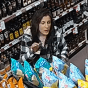Une femme debout face à la caméra de sécurité à l’intérieur du commerce. Elle a de longs cheveux brun foncé ou noirs et des ongles peints. Elle porte un manteau à carreaux noirs et blancs avec un capuchon, ainsi qu’un tee-shirt rose pâle. Au premier plan, on voit des sacs de croustilles et, en arrière-plan, des bouteilles d’alcool.