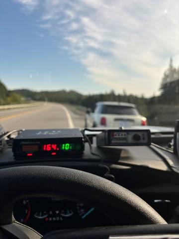 A car is pulled over on the side of a road. A police vehicle’s radar is pictured in the foreground displaying a speed of 164 km/h.