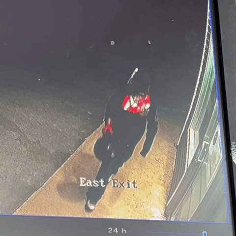 An individual with their face covered, wearing a black, red and white sweater and carrying a helmet, walks toward a storefront;