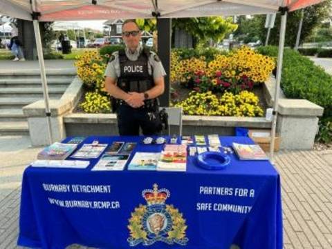 An RCMP officer in uniform stands outdoors near a flowerbed at a table with blue Burnaby RCMP tablecloth on it with a number of promotional items