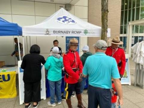 Two RCMP officers in Red Serge speak with a senior outdoors at an event near a Burnaby RCMP pop up tent