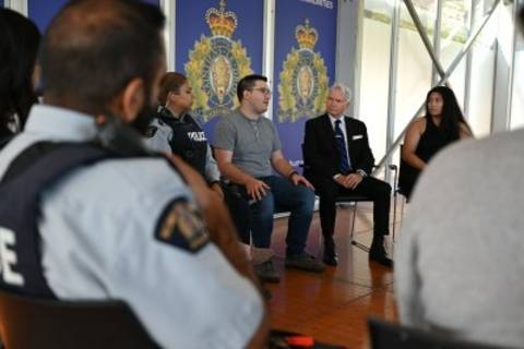 Une image montrant le dos d'un policier et plusieurs personnes en tenue civile assises sur des chaises en cercle dans un bâtiment portant les logos de la GRC.